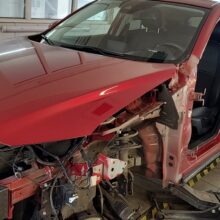 Czerwony samochód przechodzi gruntowne naprawy w garażu serwisowym, po usunięciu przednich elementów, odsłaniając wewnętrzną konstrukcję i mechanikę.