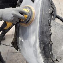 Osoba ubrana w szare rękawiczki używa szlifierki elektrycznej do polerowania zmatowiałego reflektora samochodowego. W pobliżu znajdują się narzędzia i odkurzacz, a podłoga wyłożona jest kafelkami.
