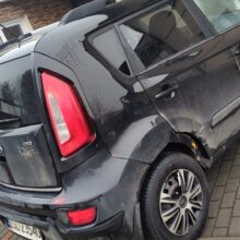 Czarna Kia Soul z widocznymi uszkodzeniami i zadrapaniami po lewej stronie jest zaparkowana na podjeździe obok domu z akcentami z cegły i kamienia. Tylne lewe koło i obszar drzwi noszą ślady uderzenia.