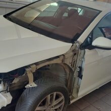 Biały samochód w garażu z usuniętym lewym przednim błotnikiem i zderzakiem, przez co widoczne są wewnętrzne elementy i lewe przednie koło.