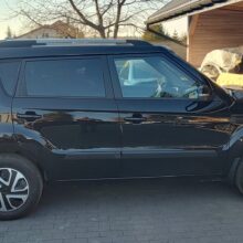 Czarny kompaktowy SUV zaparkowany na wybrukowanym podjeździe obok drewnianego garażu z innym samochodem i różnymi przedmiotami w środku. Drzewa i domy są widoczne w tle w słoneczny dzień.