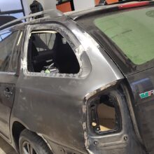 Ciemny SUV w warsztacie z brakującą tylną szybą i tylnym światłem, z widocznymi śladami naprawy karoserii, odsłoniętym metalem i śladami szlifowania. Wewnątrz pojazdu i wokół miejsca pracy widoczne są narzędzia i sprzęt.