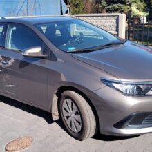 Szary samochód typu hatchback z widocznymi zarysowaniami i wgnieceniami na przednich drzwiach pasażera jest zaparkowany na wybrukowanym podjeździe w pobliżu ogrodzenia i budynków mieszkalnych.