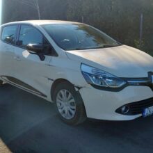 Biały Renault Clio z widocznym uszkodzeniem lewego przedniego błotnika stoi zaparkowany na nasłonecznionej ulicy, w tle widać drzewa i inne samochody.