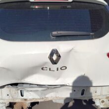 Biały samochód Renault Clio z poważnymi uszkodzeniami tylnego zderzaka i bagażnika, w tym dużym wgnieceniem i brakującą tablicą rejestracyjną, zaparkowany na zewnątrz na utwardzonej nawierzchni.