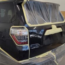 Czarny SUV zaparkowany w warsztacie, częściowo przykryty folią plastikową i taśmą maskującą, przygotowany do malowania. Okna, światła i krawędzie pojazdu są zaklejone taśmą w celu ochrony. W pobliżu widoczne są narzędzia i chłodnica.