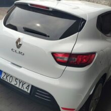 Biały Renault Clio zaparkowany na szarym ceglanym podjeździe w pobliżu zadaszonego pojazdu, z kamiennym murem i drzwiami garażowymi w tle. Na tablicy rejestracyjnej samochodu widnieje napis „2KO SYK4.