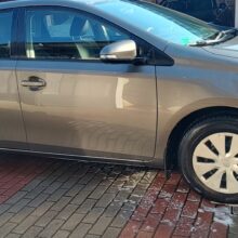 Czysty, jasnobrązowy samochód typu hatchback zaparkowany na wybrukowanym podjeździe. W pobliżu opon widać pianę mydlaną, co wskazuje, że samochód został właśnie umyty. W tle widać inne pojazdy i garaż.