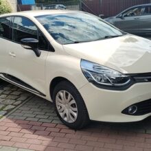 Biały hatchback Renault Clio jest zaparkowany na wybrukowanym podjeździe z szarym samochodem i budynkami w tle. Samochód jest zwrócony lekko w lewo, a światło słoneczne odbija się od jego powierzchni.