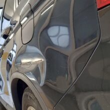 Zbliżenie na boczny widok ciemnego samochodu z dużym wgnieceniem i zadrapaniami nad tylnym kołem, zaparkowanego w garażu, z innym samochodem widocznym przez okno.