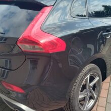 Czarny samochód Volvo hatchback zaparkowany na utwardzonym terenie obok beżowego pojazdu. Widoczny jest tył i bok samochodu, z odbiciami drzew na powierzchni i europejskimi tablicami rejestracyjnymi.