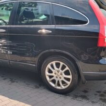 Czarny SUV zaparkowany na wybrukowanym podjeździe obok brązowego pojazdu. Na zdjęciu widać bok samochodu, podkreślając jego czyste i lśniące nadwozie. Scena rozgrywa się na zewnątrz w słoneczny dzień.