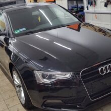 Czarny sedan Audi stoi zaparkowany w czystym, dobrze oświetlonym garażu z kafelkowymi ścianami, narzędziami zawieszonymi po prawej stronie i dużymi czarnymi drzwiami w tle.