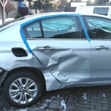 Srebrny sedan ze znacznymi uszkodzeniami lewej strony, w tym wgniecionymi tylnymi drzwiami, brakującą pokrywą korka wlewu paliwa i zniszczonym tylnym zderzakiem, jest zaparkowany na asfaltowym podjeździe w pobliżu innych pojazdów i budynków.