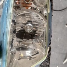 Zbliżenie reflektora samochodowego zamaskowanego taśmą i plastikiem, prawdopodobnie przygotowanego do malowania lub naprawy w warsztacie samochodowym. Reflektor jest błyszczący i czysty, otoczony owiniętymi prętami i płytkami podłogowymi.